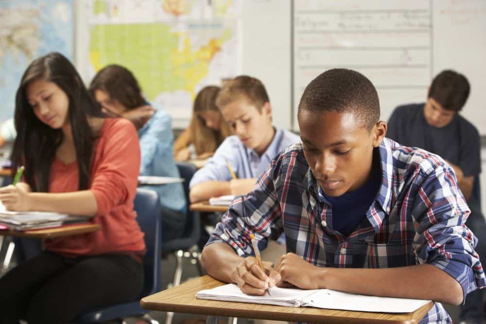 Teenage African American male wearing a plaid button-up shirt sitting at a desk in class. He is writing in a notebook on his desk using a pencil. Around him are his classmates, also writing on their papers. They are all concentrating on their own work.